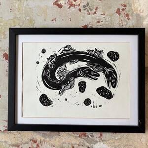 K&ouml;nnte beinhalten: Schwarz-Wei&szlig;-Linolschnitt mit zwei schwimmenden Fischen, umgeben von kleineren Formen. Das Kunstwerk ist in einem schwarzen Rahmen mit wei&szlig;em Passepartout gerahmt. Der Druck wird vor einer strukturierten, gealterten Wand pr&auml;sentiert.