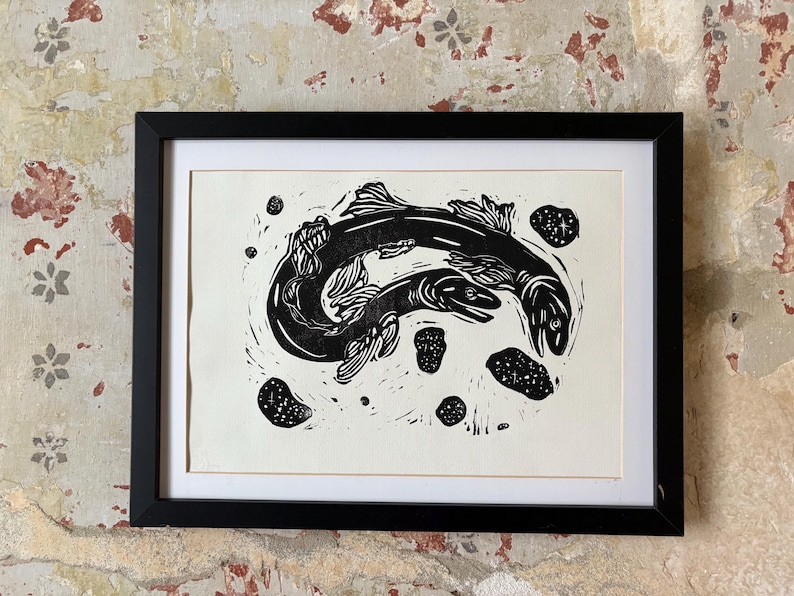 K&ouml;nnte beinhalten: Schwarz-Wei&szlig;-Linolschnitt mit zwei schwimmenden Fischen, umgeben von kleineren Formen. Das Kunstwerk ist in einem schwarzen Rahmen mit wei&szlig;em Passepartout gerahmt. Der Druck wird vor einer strukturierten, gealterten Wand pr&auml;sentiert.