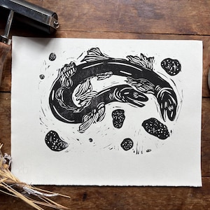 K&ouml;nnte beinhalten: Schwarz-Wei&szlig;-Linolschnitt mit zwei schwimmenden Fischen und mehreren kleinen, gesprenkelten Formen. Der Druck befindet sich auf wei&szlig;em Papier, mit einer Holzwalze und einem Farbbeh&auml;lter im Hintergrund. Auf einer Holzoberfl&auml;che.