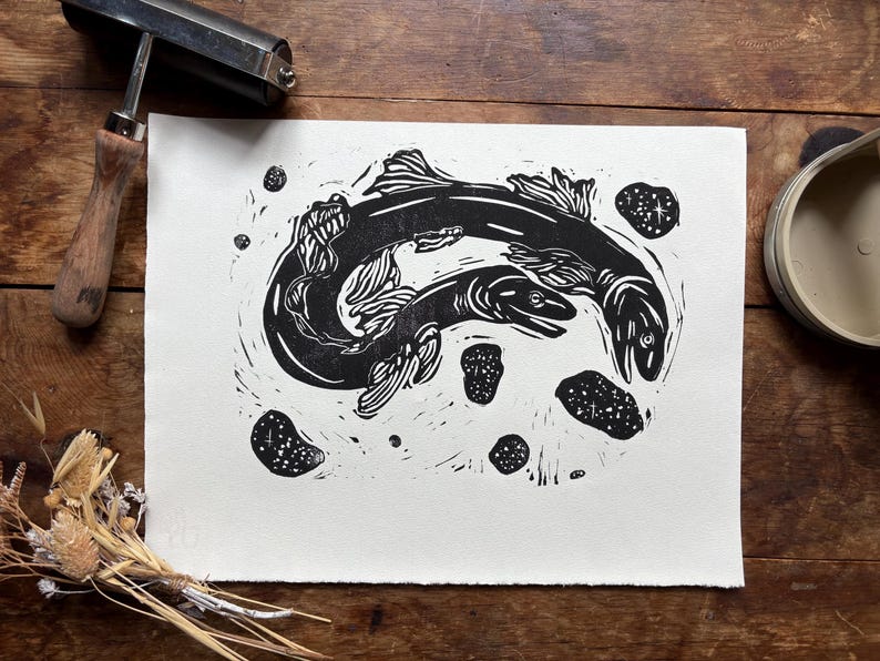 K&ouml;nnte beinhalten: Schwarz-Wei&szlig;-Linolschnitt mit zwei schwimmenden Fischen und mehreren kleinen, gesprenkelten Formen. Der Druck befindet sich auf wei&szlig;em Papier, mit einer Holzwalze und einem Farbbeh&auml;lter im Hintergrund. Auf einer Holzoberfl&auml;che.