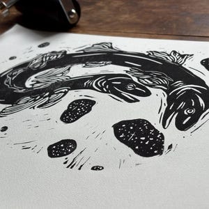 K&ouml;nnte beinhalten: Ein schwarz-wei&szlig; Linolschnitt mit zwei Fischen, die sich kreisf&ouml;rmig bewegen. Die Fische sind detailliert mit Flossen und Schuppen dargestellt und von kleinen, gepunkteten Formen umgeben. Der Druck ist auf wei&szlig;em Papier.