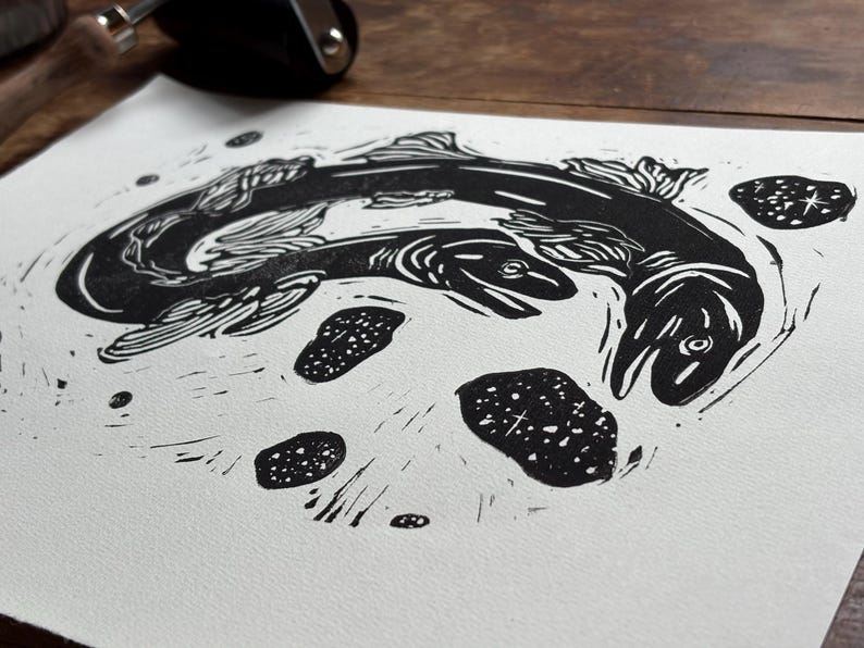 K&ouml;nnte beinhalten: Ein schwarz-wei&szlig; Linolschnitt mit zwei Fischen, die sich kreisf&ouml;rmig bewegen. Die Fische sind detailliert mit Flossen und Schuppen dargestellt und von kleinen, gepunkteten Formen umgeben. Der Druck ist auf wei&szlig;em Papier.