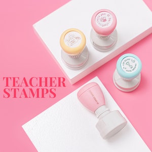 May include: Teacher stamps with pastel-coloured tops and motivational messages. The stamps have a clear base and are arranged on a white surface against a pink background. The text "TEACHER STAMPS" is displayed in pink.