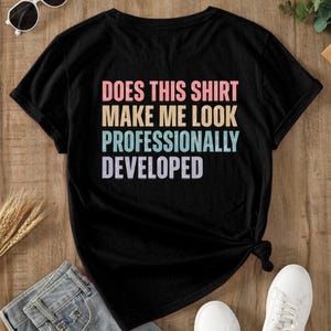May include: A black short-sleeved t-shirt featuring the text "DOES THIS SHIRT MAKE ME LOOK PROFESSIONALLY DEVELOPED" printed in a stacked, multi-colored retro font with pink, yellow, teal, and light purple lettering against the dark fabric.