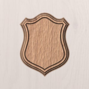 Puede incluir: Placa de madera en forma de escudo con superficie texturizada. La placa es de color marrón claro con ribetes marrones más oscuros que delinean los bordes. El escudo tiene una parte superior decorativa y festoneada.