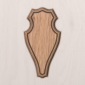 May include: A wooden plaque with a shield shape, featuring a textured surface and a dark border. The plaque has a carved design and is set against a light background. It is likely used for display or decoration.