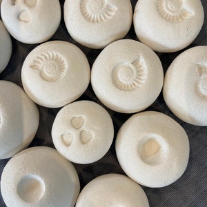 May include: A collection of white, round, ceramic stones with various designs, including hearts, spirals, and circles. The stones are smooth and have a matte finish.