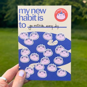 以下が含まれることがあります： クリーム色の背景に青い文字で my new habit is to go outside every day と書かれた長方形の習慣トラッカーカード。テキストの下には、王冠をかぶった小さな薄ピンク色のカエルのイラストが青い池の背景に多数描かれています。