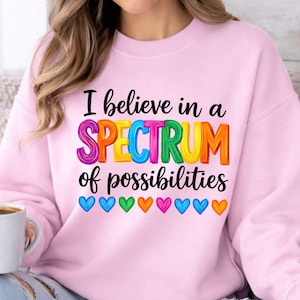May include: A light pink sweatshirt with the text "I believe in a SPECTRUM of possibilities" in colorful letters. Below the text are colorful heart graphics. The sweatshirt is a digital product.