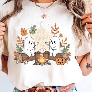 May include: Cream-colored t-shirt featuring a Halloween-themed graphic. Two cartoon ghosts roast marshmallows over a campfire, with a pumpkin, mushrooms, and fall foliage. The design is centered on the shirt.