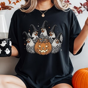 May include: Black t-shirt featuring a Halloween design. The graphic shows five chickens wearing witch hats around a carved pumpkin. A black mug with white ghost designs is held in the left hand. An orange pumpkin sits on the right.