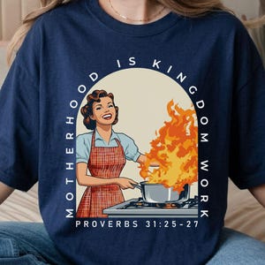 Puede incluir: Camiseta azul marino con una ilustración retro de una mujer cocinando con llamas saliendo de una olla. El texto dice "Motherhood is Kingdom Work" y "Proverbios 31:25-27" en un arco blanco. La camiseta es de estilo casual y cómodo.