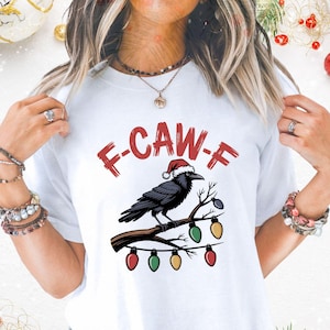 Può includere: T-shirt bianca con grafica di un corvo nero con cappello di Babbo Natale appollaiato su un ramo con luci natalizie. Il testo "F-CAW-F" è sopra il corvo in lettere rosse. Abbigliamento festivo natalizio.