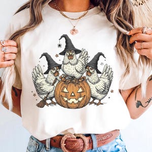 May include: Cream-colored t-shirt featuring a Halloween-themed graphic. The design shows three white chickens wearing black witch hats, gathered around a carved orange pumpkin. A spider is on one hat. Stars and a broom complete the design.