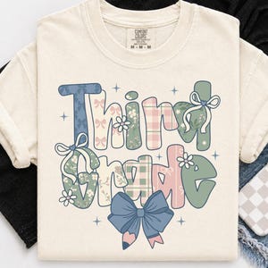 Puede incluir: Una camiseta de color crema con el texto Third Grade en letras grandes y estilizadas. Las letras están rellenas con patrones de lazos azules, cuadros rosas y estampados florales verdes. Un lazo azul grande está centrado en la parte inferior.