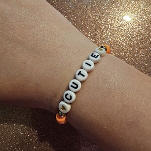 Op de afbeelding: Een witte kralen armband met het woord "CUTIE" gespeld in zwarte letters. De armband heeft een kleine gouden hart charme en oranje kralen.