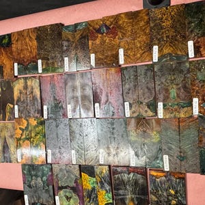May include: A selection of rectangular wooden pieces, each displaying unique colours and patterns. The wood grain is visible, with hues of brown, gold, green, and grey. Small white labels are attached to each piece, likely for identification.