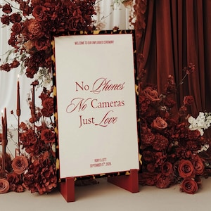 Puede incluir: Un letrero de bienvenida para una ceremonia sin teléfonos, con el texto "No Phones, No Cameras, Just Love". El letrero está rodeado de rosas burdeos, velas y arreglos florales. El letrero está sobre un caballete rojo.