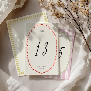 Pode incluir: Um cartão de número de mesa branco com uma borda vermelha festoneada. O número 13 está escrito em fonte cursiva preta. O texto "TABLE" está impresso em preto no topo do cartão. O texto "MARY & GREGG 05.01.26" está impresso em preto na parte inferior do cartão.