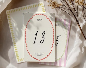 May include: A white table number card with a red scalloped border. The number 13 is written in black cursive font. The text "TABLE" is printed in black at the top of the card. The text "MARY & GREGG 05.01.26" is printed in black at the bottom of the card.