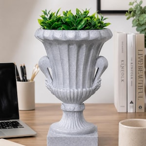 Op de afbeelding: Een grijze, sierlijke urnvormige plantenbak met een vierkante basis, gevuld met levendig groen kunstmatig gebladerte. De plantenbak heeft decoratieve handvatten en verticale groeven. Het staat op een houten oppervlak, wat duidt op een decoratief woonaccessoire.