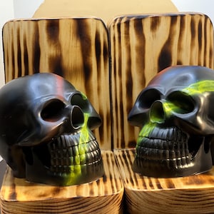 May include: Two black skull sculptures with green and red accents. The skulls are displayed on wooden pedestals with a burnt wood design. The skulls have detailed facial features and are positioned facing each other.
