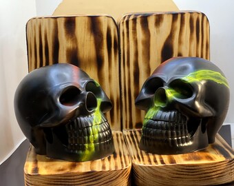 May include: Two black skull sculptures with green and red accents. The skulls are displayed on wooden pedestals with a burnt wood design. The skulls have detailed facial features and are positioned facing each other.