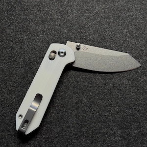 Puede incluir: Un cuchillo de bolsillo blanco con una hoja parcialmente abierta. La hoja está hecha de metal gris y tiene un acabado texturizado. El mango está hecho de un material blanco liso y tiene un pequeño botón plateado en el lateral.