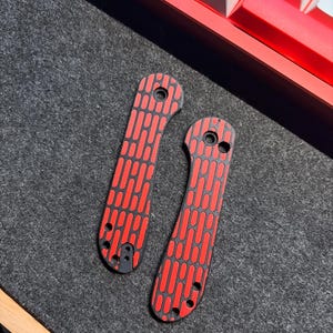 May include: Two red and black knife scales with a repeating pattern of vertical rectangles. The scales are lying on a dark gray surface, with a red keyboard in the background.