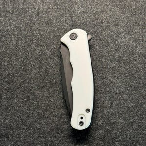 May include: A white and black pocket knife with a black blade and a white handle. The handle has a small circular indentation and two small screws.