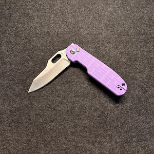May include: A folding pocket knife with a silver-toned metal blade and a light purple handle. The handle features a textured, brick-like pattern and a silver pivot screw. The blade has a thumb hole cutout near the base for opening the mechanism.