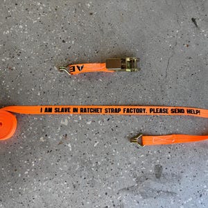 May include: Orange ratchet strap with a black printed message "I AM SLAVE IN RATCHET STRAP FACTORY. PLEASE SEND HELP!" and a gold metal buckle.