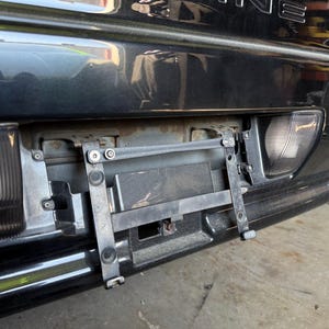 May include: Close-up of a black car's rear bumper, showcasing a license plate bracket and taillight. The bumper has a glossy finish, with a metal bracket attached. The taillight is red and clear. The car appears to be in a garage.