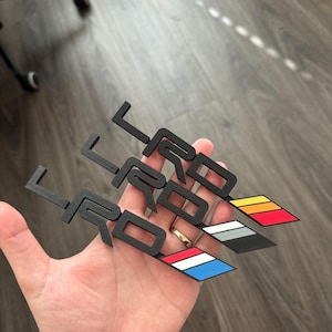 以下が含まれることがあります： スタイリッシュなフォントで「TRD」と書かれた、黒い金属製の車のエンブレムが3つ。各エンブレムには、赤、白、青、グレー、白、オレンジ、黒、白、黄色の異なる色のストライプデザインが施されています。