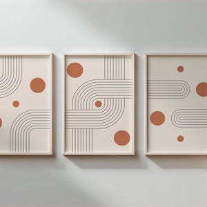 Puede incluir: Tres impresiones artísticas abstractas enmarcadas. Cada impresión presenta un fondo crema con líneas curvas negras y círculos color óxido. Los marcos son blancos. El arte se exhibe en una pared gris claro.