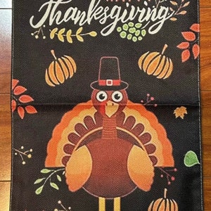 May include: A black and orange Thanksgiving garden flag with a cartoon turkey wearing a pilgrim hat and the text "Happy Thanksgiving".