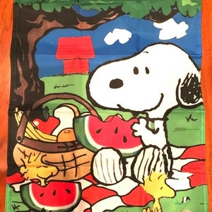 May include: Snoopy and Woodstock enjoy a picnic under a tree. Snoopy is holding a slice of watermelon and Woodstock is eating a piece of bread. A red doghouse is in the background.