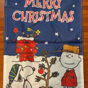 May include: A blue Christmas flag featuring Snoopy, Charlie Brown, and a Christmas tree. The flag has the words "MERRY CHRISTMAS" in red and white. Snoopy is wearing a winter hat, and Charlie Brown is wearing a red coat.