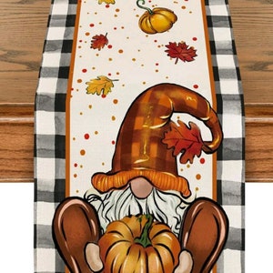 May include: A white table runner with a black and white checkered border. The center of the runner features a gnome holding a pumpkin. The gnome is wearing a brown hat with a plaid pattern and has a white beard. The background is white with orange and brown leaves and orange dots.