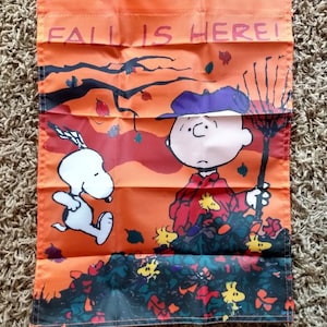 May include: An orange and black garden flag with the text "Fall is Here!"  Snoopy the beagle is running to the right and Charlie Brown is raking leaves.  There are small yellow birds and red and orange leaves scattered around them.