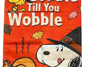 May include: Thanksgiving-themed garden flag with Snoopy and Woodstock characters. The flag is orange with the text "Gobble Till You Wobble" and illustrations of a turkey and autumn leaves. The Snoopy character is wearing a pilgrim hat.