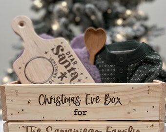 Op de afbeelding: Een houten doos met de tekst "Christmas Eve Box for The Samaniego Family" erop gegraveerd. De doos is versierd met sterren, harten en kerstboompjes.