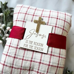 May include: A red and white plaid blanket wrapped with a red ribbon and a wooden sign that reads "Jesus is the reason for the season". A gold glitter cross is attached to the sign.