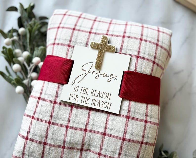 May include: A red and white plaid blanket wrapped with a red ribbon and a wooden sign that reads "Jesus is the reason for the season". A gold glitter cross is attached to the sign.