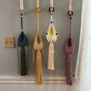 May include: Four macrame plant hangers with wooden dowels and beaded accents. The hangers are in various colors, including green, beige, white, and pink.