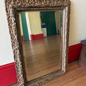 May include: A rectangular wall mirror featuring a thick, ornate gold-toned frame with intricate scrollwork and relief detailing. The glass surface is clear and reflective, showing a room interior with wooden flooring and red baseboards.