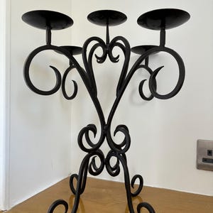 May include: A black metal candelabra featuring five circular candle plates arranged at varying heights. The structure is composed of curved, scroll-like metal rods that form an intricate, symmetrical base and branching arms for holding candles.
