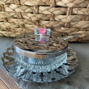 May include: A clear glass sugar bowl with a silver lid and a decorative silver base. The lid has a small, square, clear handle with a pink and blue floral design. The bowl sits on a silver, scalloped edge plate.