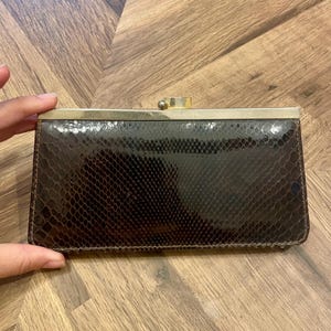 May include: A rectangular clutch bag featuring a dark brown snakeskin-textured exterior with a glossy finish. The bag is topped with a metallic gold-toned frame and a central spherical clasp closure, providing a structured and sleek appearance.
