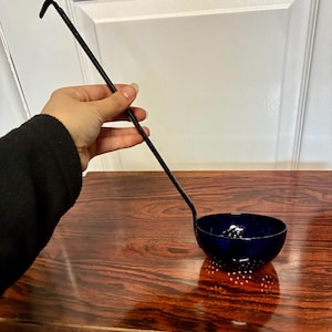 May include: A dark blue metal skimmer ladle with a long, slender black handle. The bowl of the ladle features a series of small, evenly spaced circular perforations throughout its base and sides, designed for straining liquids from solid food items.